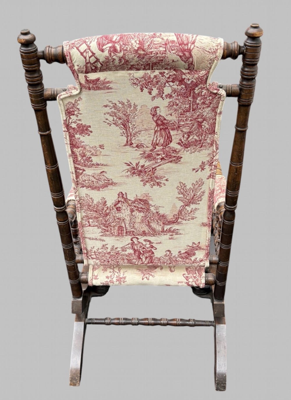 An antique turned wood rocking chair featuring dark timber spindle armrests and frame with cream fabric upholstery displaying red figurative pattern work. View IV
