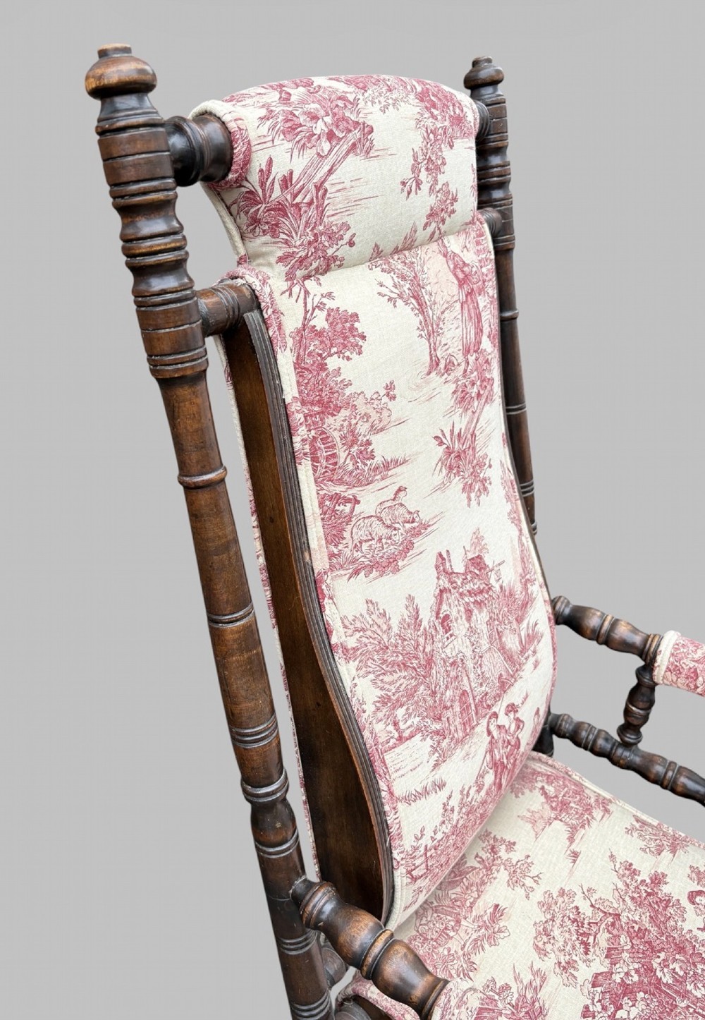 An antique turned wood rocking chair featuring dark timber spindle armrests and frame with cream fabric upholstery displaying red figurative pattern work. View VI