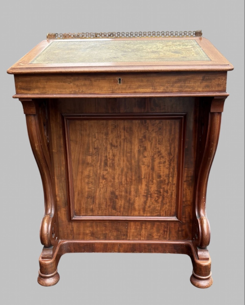 A front view of a Victorian walnut Davenport desk featuring a slanted green leather writing surface, decorative top brass gallery rail, carved front supports, and a panelled wooden body.