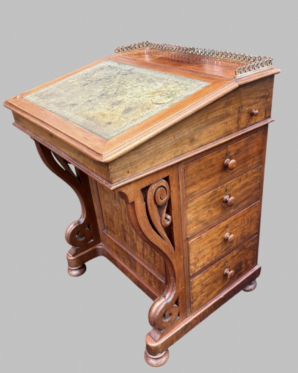 A front view of a Victorian walnut Davenport desk featuring a slanted green leather writing surface, decorative top brass gallery rail, carved front supports, and a panelled wooden body. View II