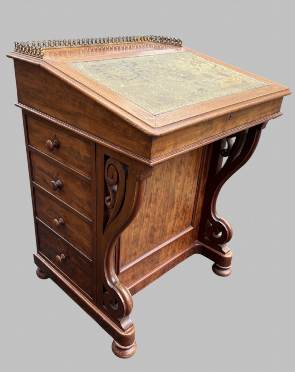 A front view of a Victorian walnut Davenport desk featuring a slanted green leather writing surface, decorative top brass gallery rail, carved front supports, and a panelled wooden body. View III