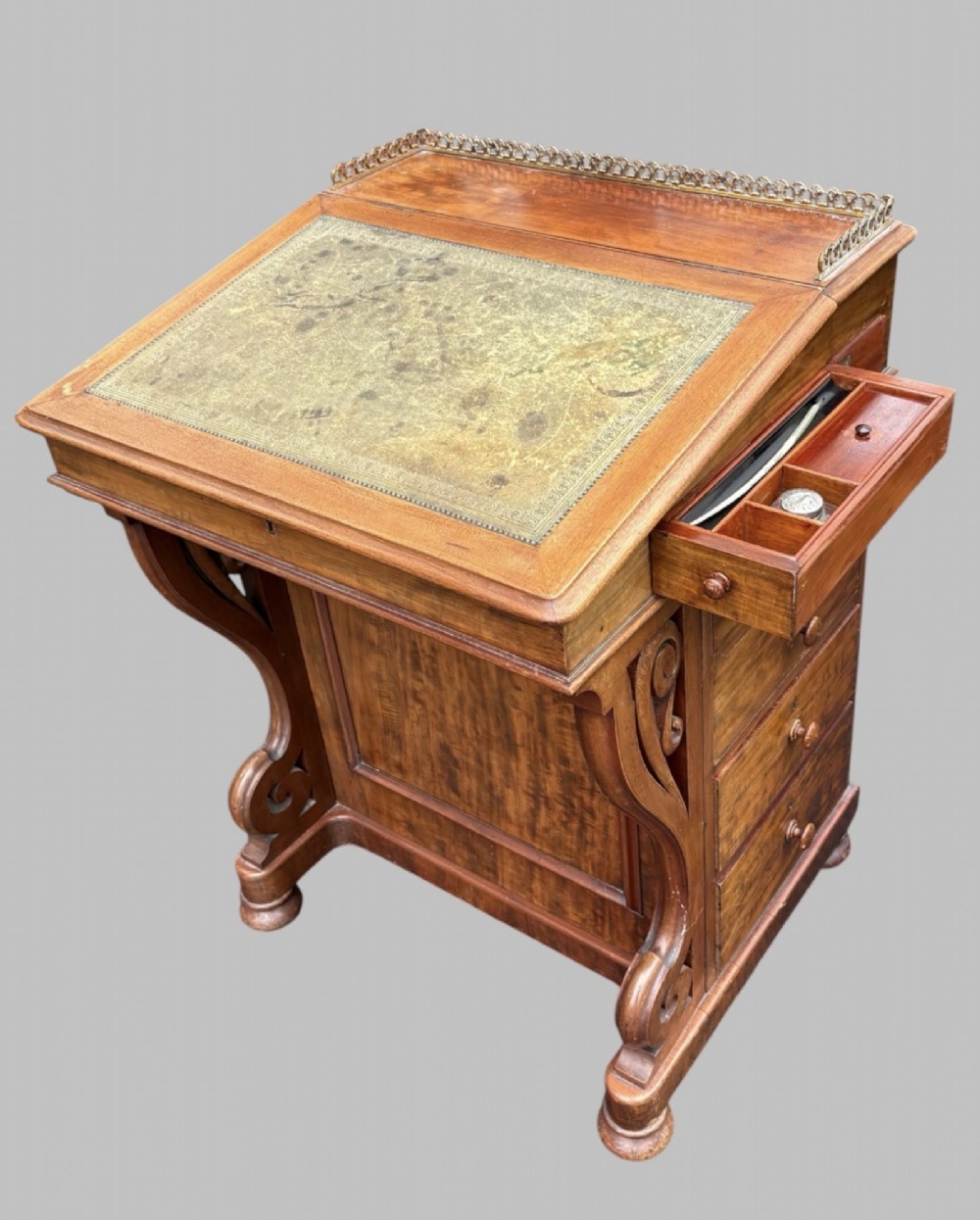 A front view of a Victorian walnut Davenport desk featuring a slanted green leather writing surface, decorative top brass gallery rail, carved front supports, and a panelled wooden body. View VI
