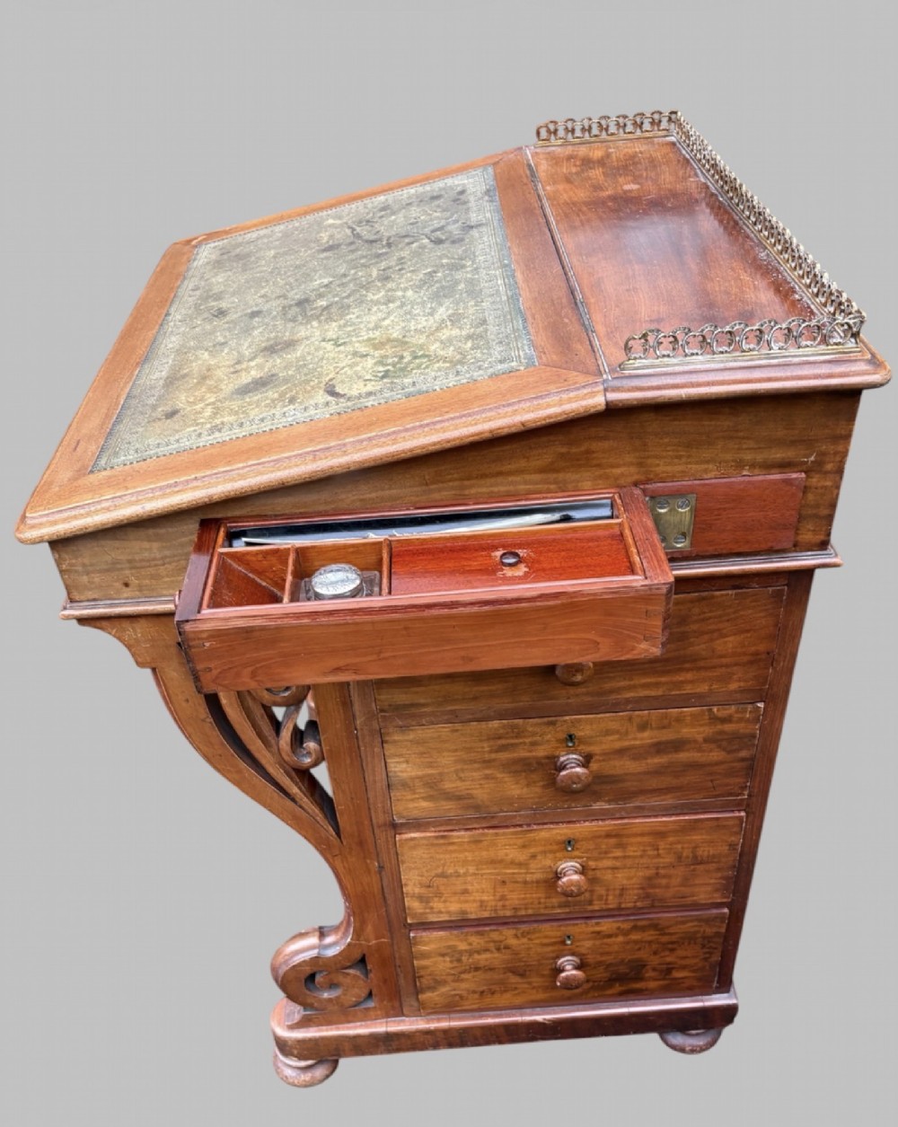 A front view of a Victorian walnut Davenport desk featuring a slanted green leather writing surface, decorative top brass gallery rail, carved front supports, and a panelled wooden body. View VII