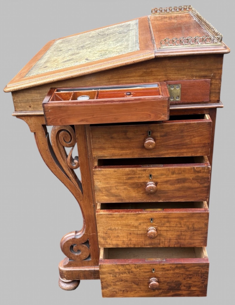 A front view of a Victorian walnut Davenport desk featuring a slanted green leather writing surface, decorative top brass gallery rail, carved front supports, and a panelled wooden body. View VIII