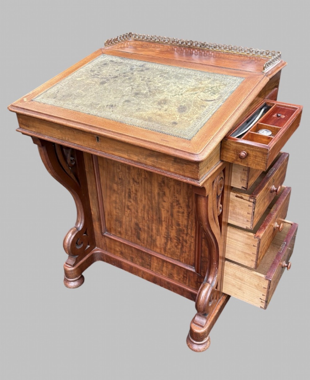 A front view of a Victorian walnut Davenport desk featuring a slanted green leather writing surface, decorative top brass gallery rail, carved front supports, and a panelled wooden body. View IX