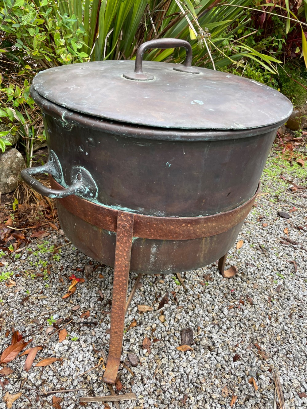 A large lidded copper log bin featuring riveted side handles, a flat lid with a central handle, and a raised iron support stand.