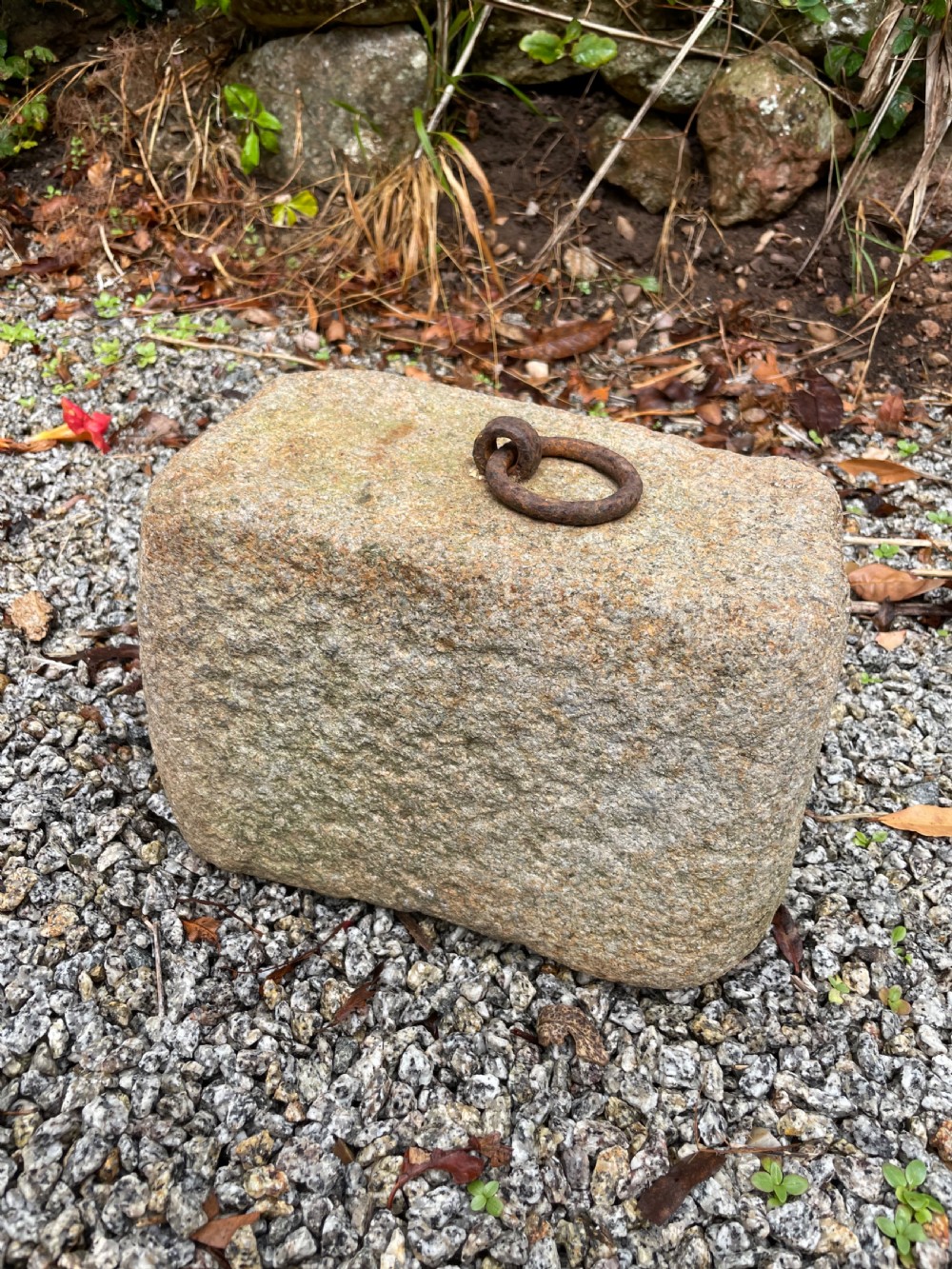 A rectangular, weathered grey granite block rests on gravel, featuring a rusted iron ring embedded in its top surface.