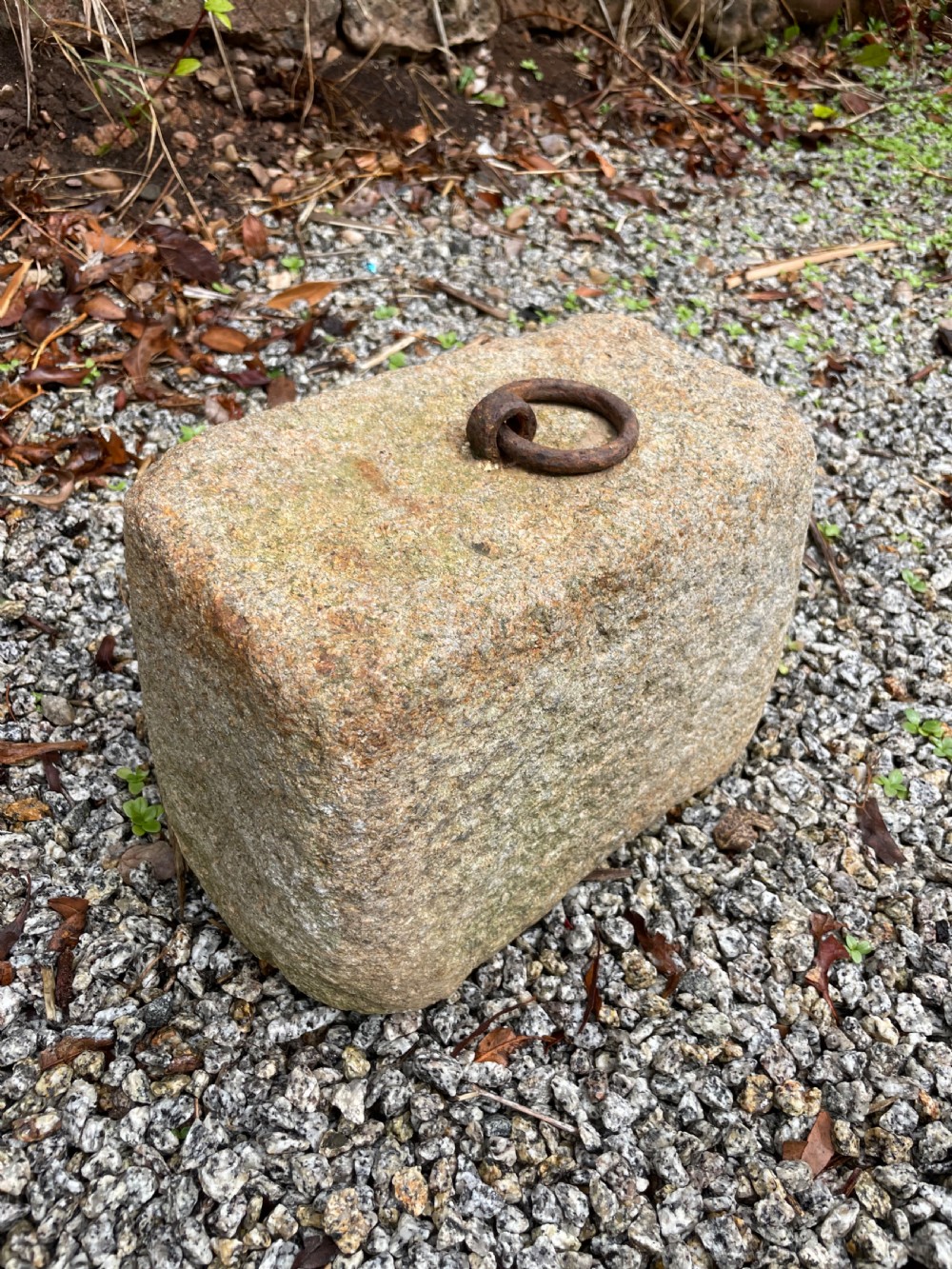 A rectangular, weathered grey granite block rests on gravel, featuring a rusted iron ring embedded in its top surface. View II