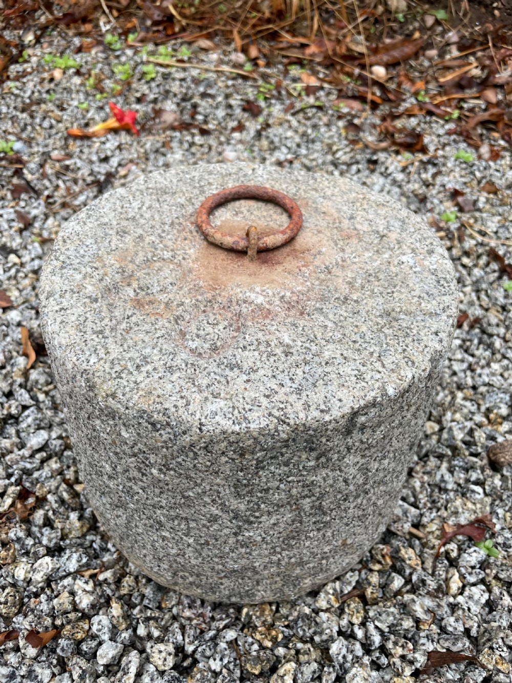A cylindrical antique granite tethering block featuring a rusted iron ring set into the top centre surface.