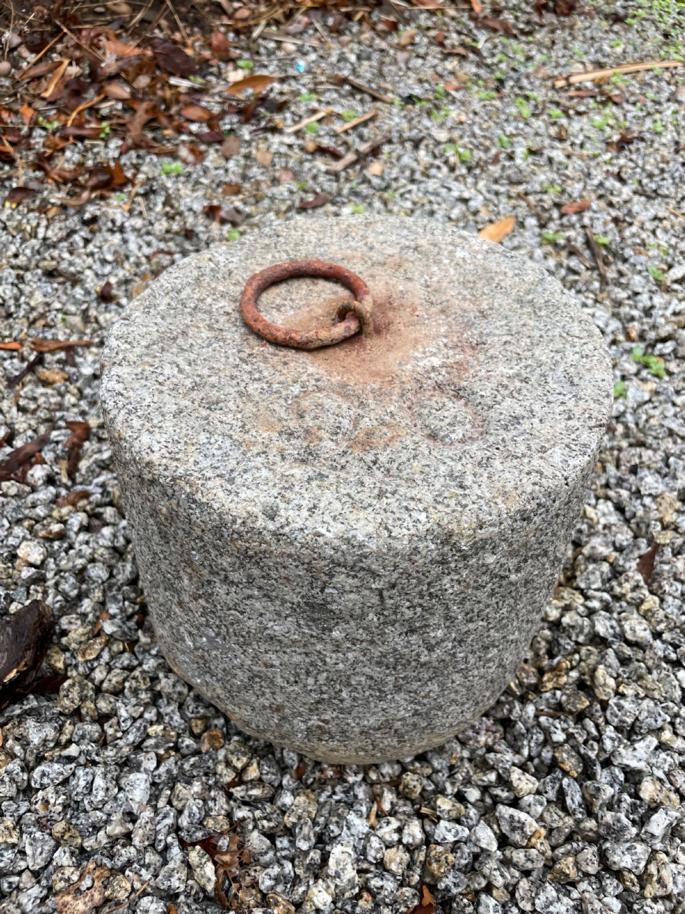 A cylindrical antique granite tethering block featuring a rusted iron ring set into the top centre surface. View IV