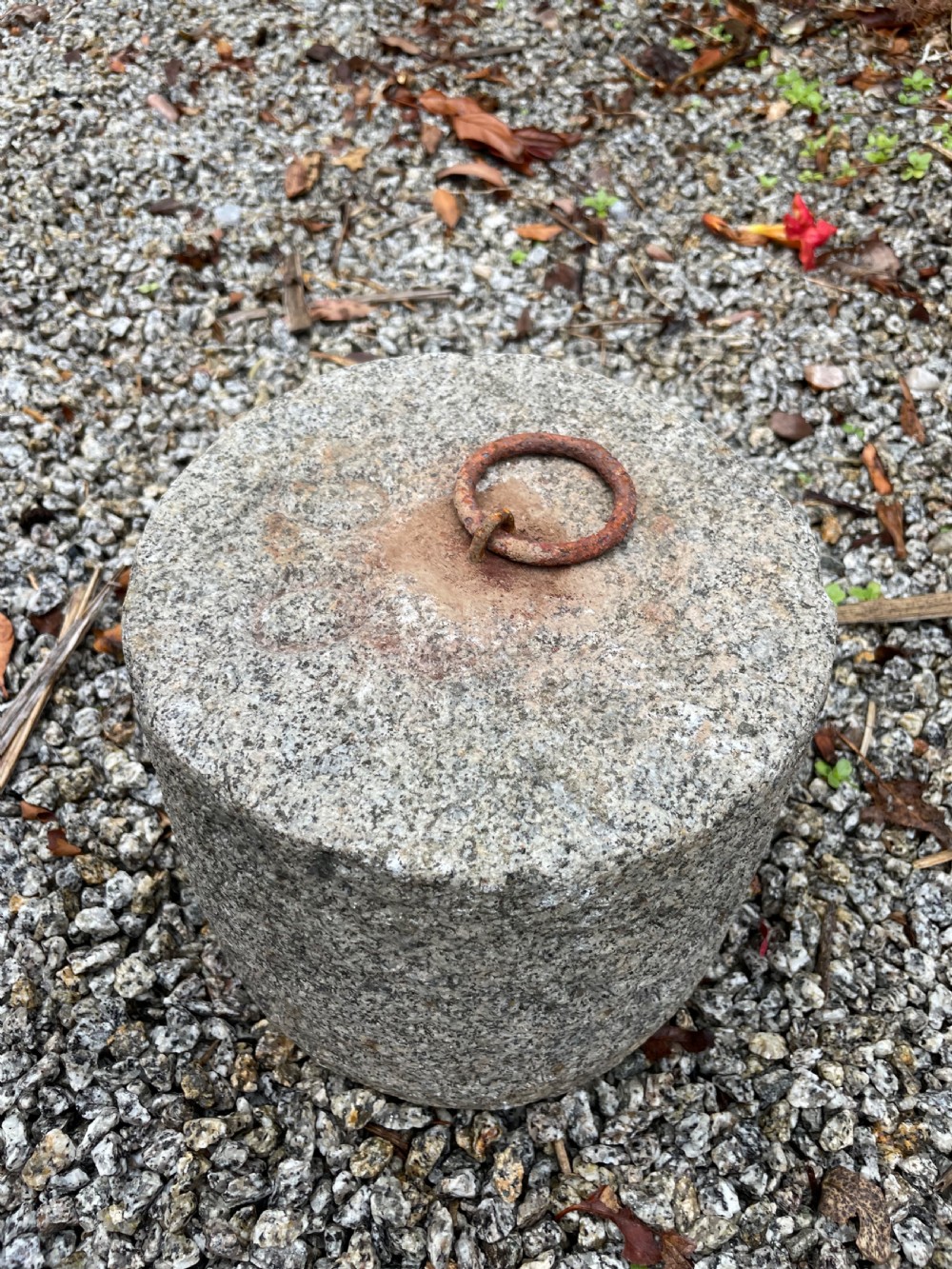 A cylindrical antique granite tethering block featuring a rusted iron ring set into the top centre surface. View V