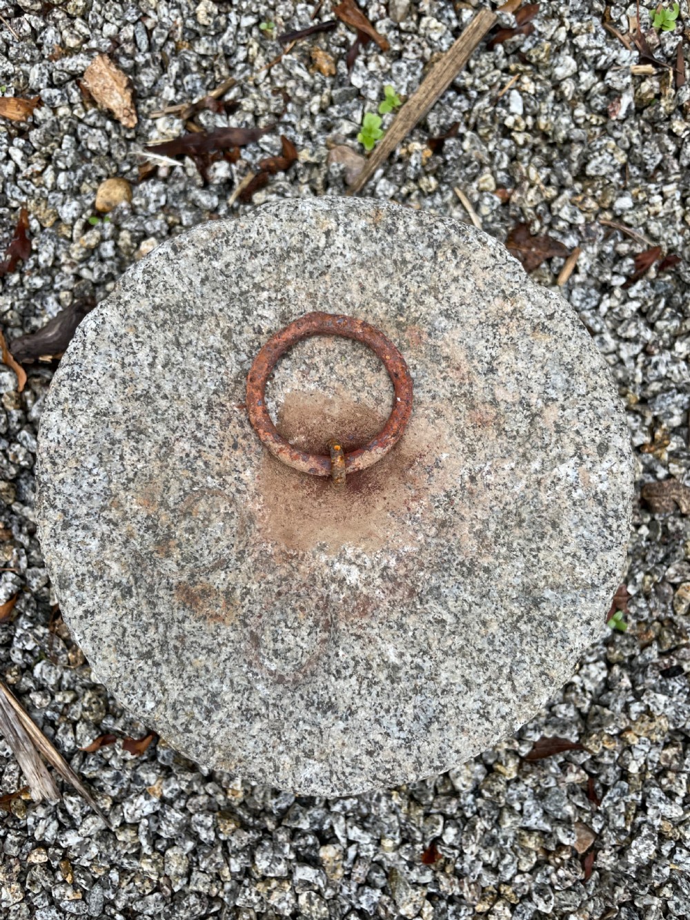 A cylindrical antique granite tethering block featuring a rusted iron ring set into the top centre surface. View VI