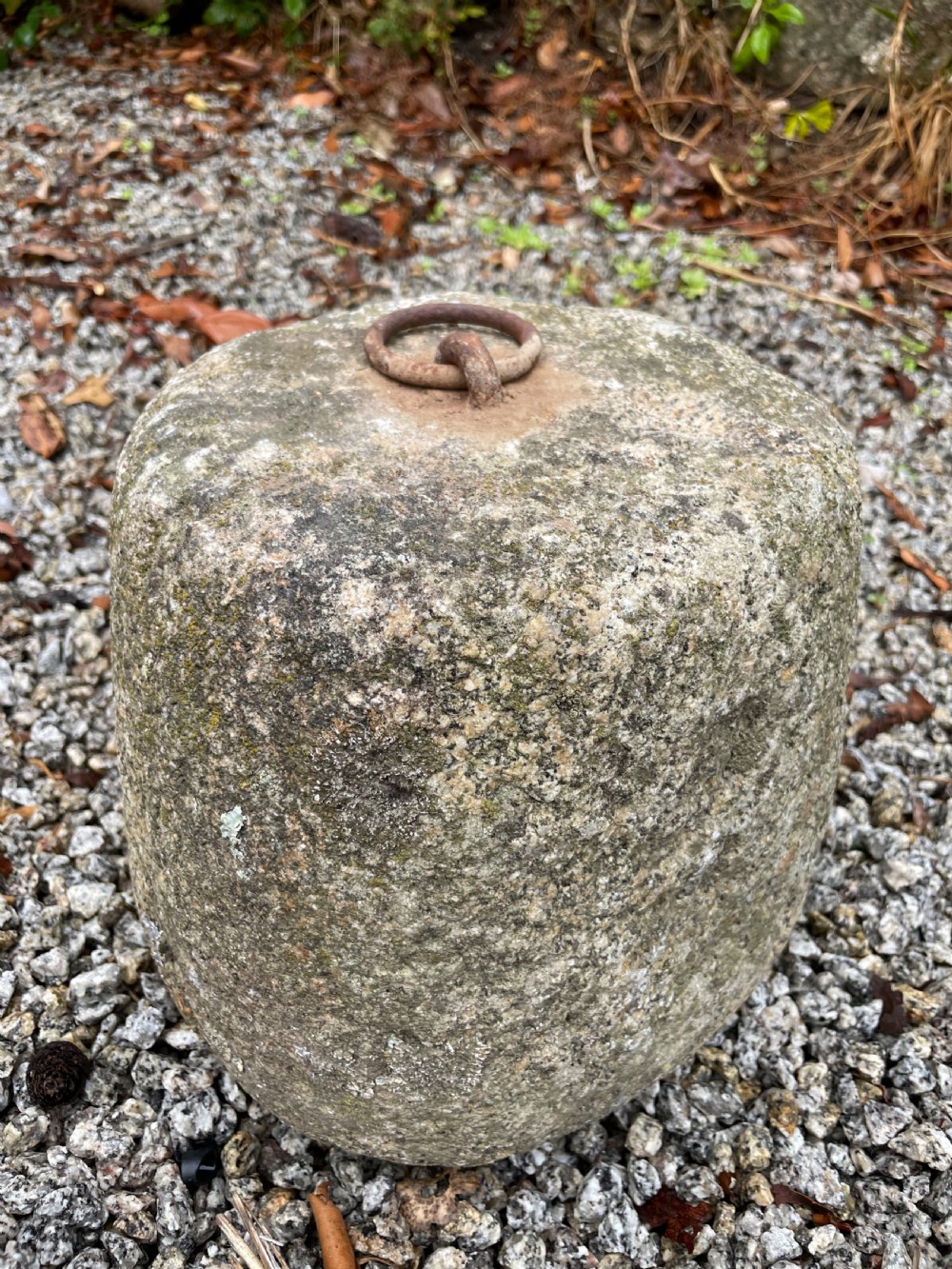 A weathered grey granite tethering block with an embedded rusted iron ring on top, resting on gravel.