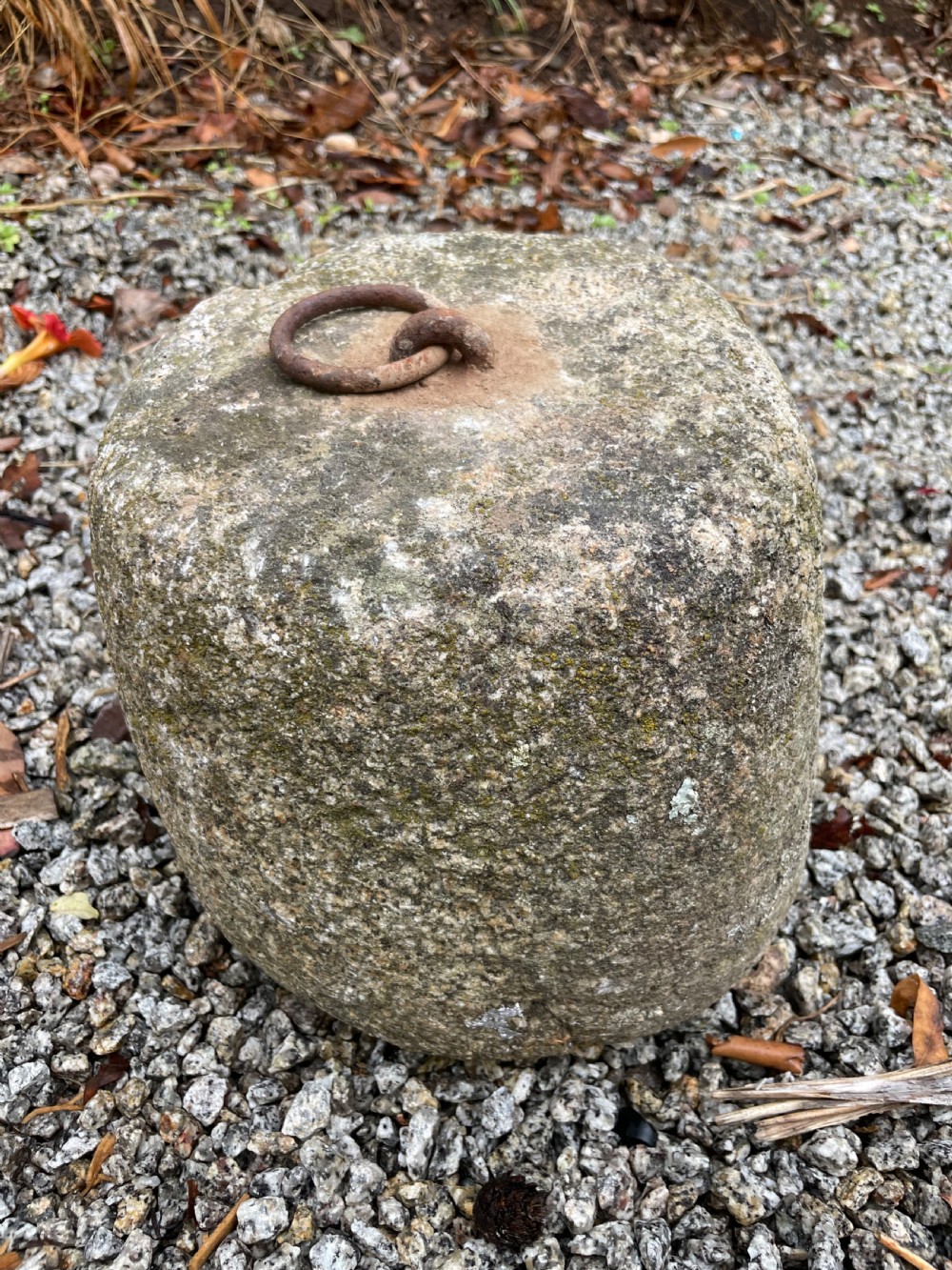 A weathered grey granite tethering block with an embedded rusted iron ring on top, resting on gravel. View II