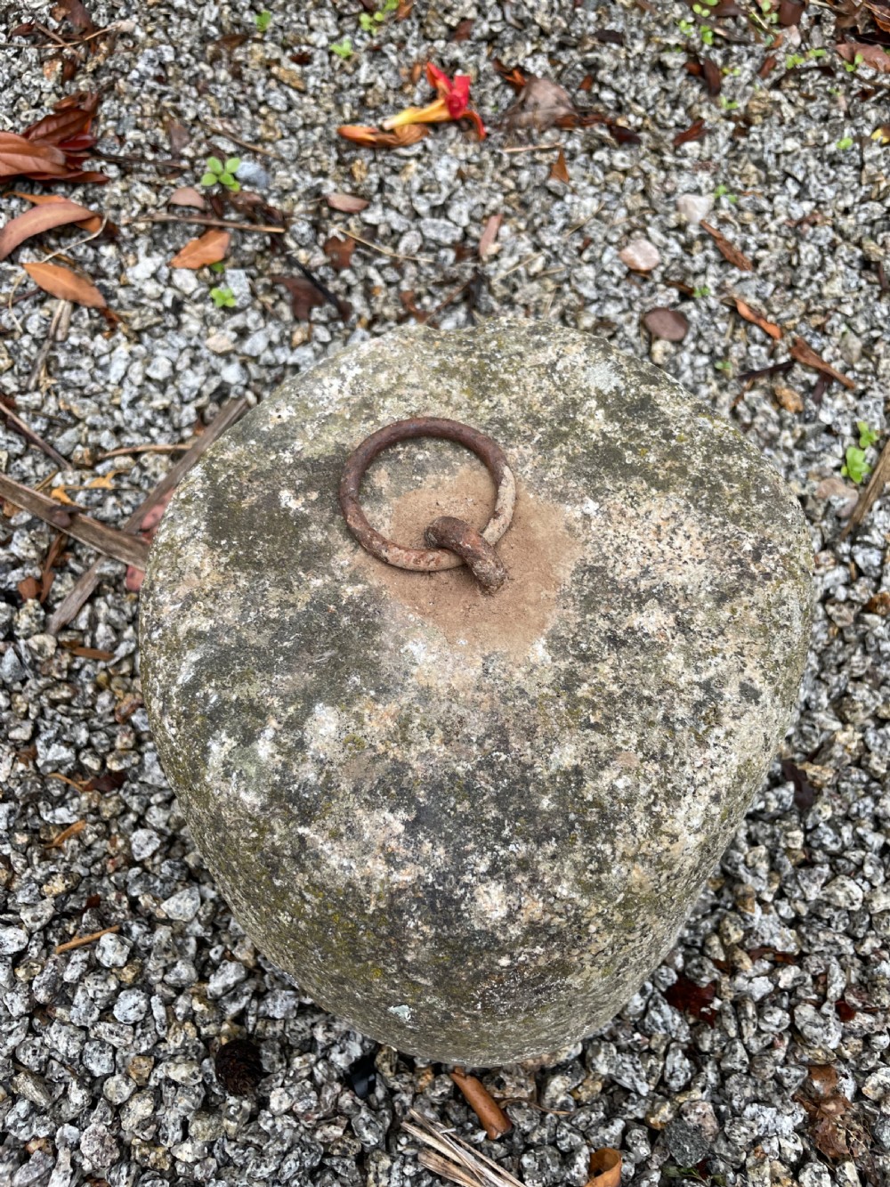 A weathered grey granite tethering block with an embedded rusted iron ring on top, resting on gravel. View III