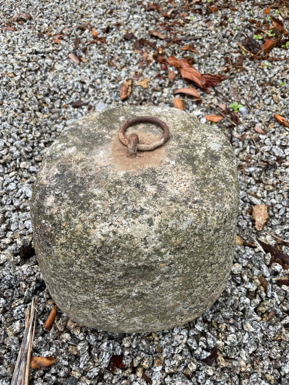 A weathered grey granite tethering block with an embedded rusted iron ring on top, resting on gravel. View IV