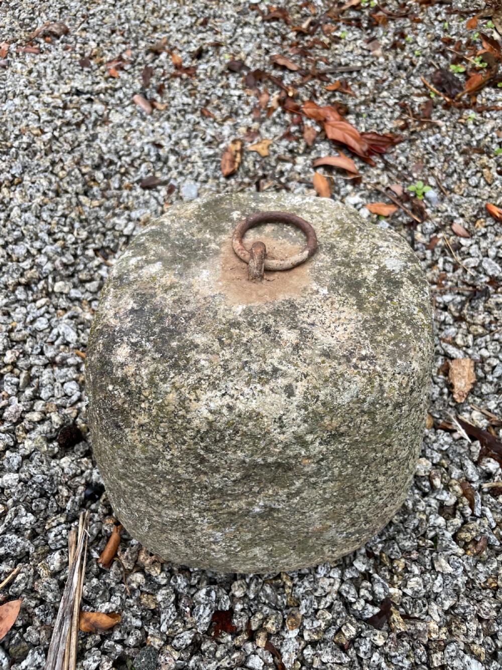 A weathered grey granite tethering block with an embedded rusted iron ring on top, resting on gravel. View V