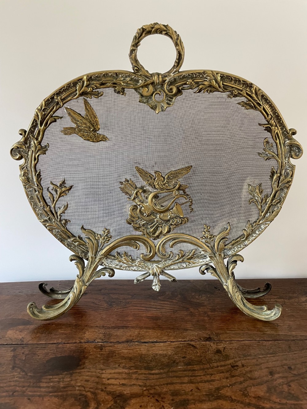 An ornate antique brass fire screen featuring pierced scrollwork, a central lyre and bird motif, and a wire mesh panel.