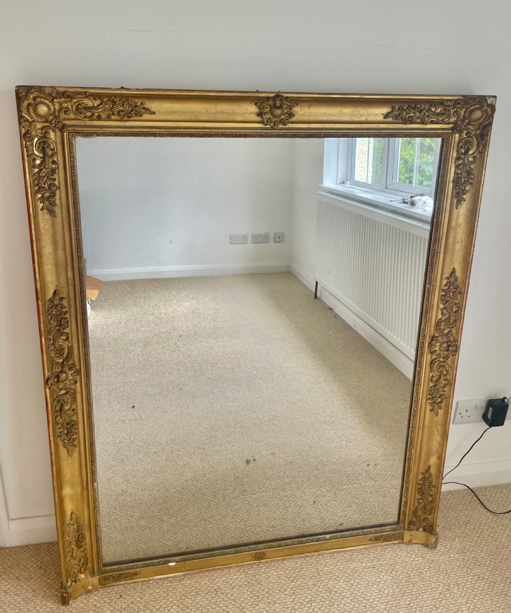 A large rectangular antique overmantel mirror featuring a gilded frame embellished with gesso scrollwork, floral motifs, and a central cartouche top emblem.