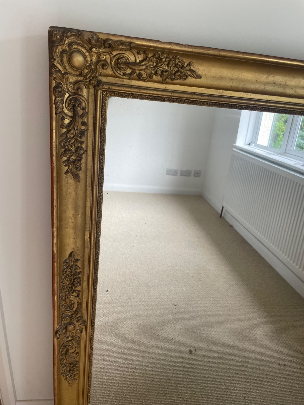 A large rectangular antique overmantel mirror featuring a gilded frame embellished with gesso scrollwork, floral motifs, and a central cartouche top emblem. View II