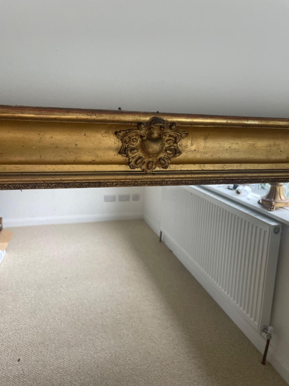 A large rectangular antique overmantel mirror featuring a gilded frame embellished with gesso scrollwork, floral motifs, and a central cartouche top emblem. View III