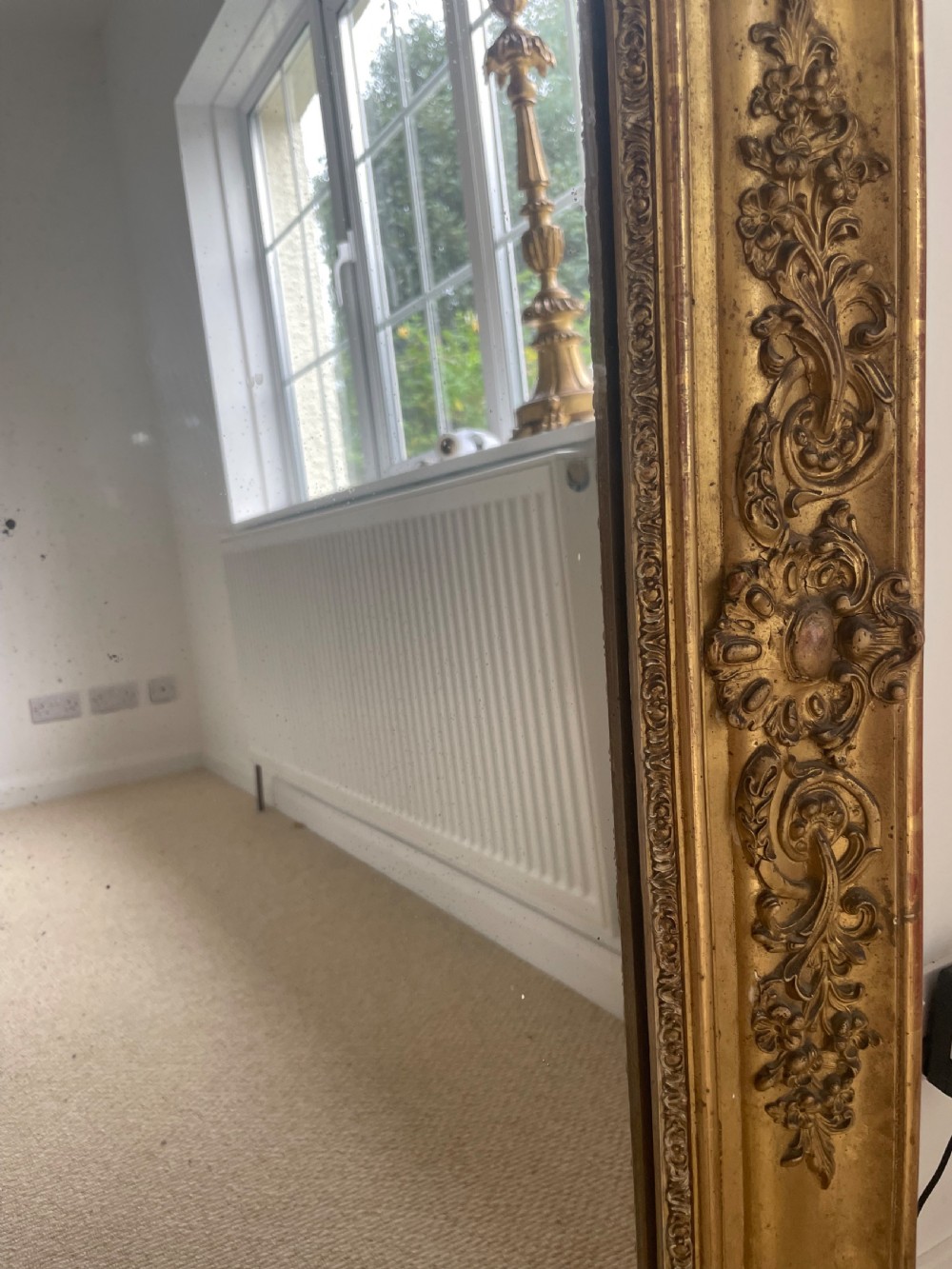 A large rectangular antique overmantel mirror featuring a gilded frame embellished with gesso scrollwork, floral motifs, and a central cartouche top emblem. View V