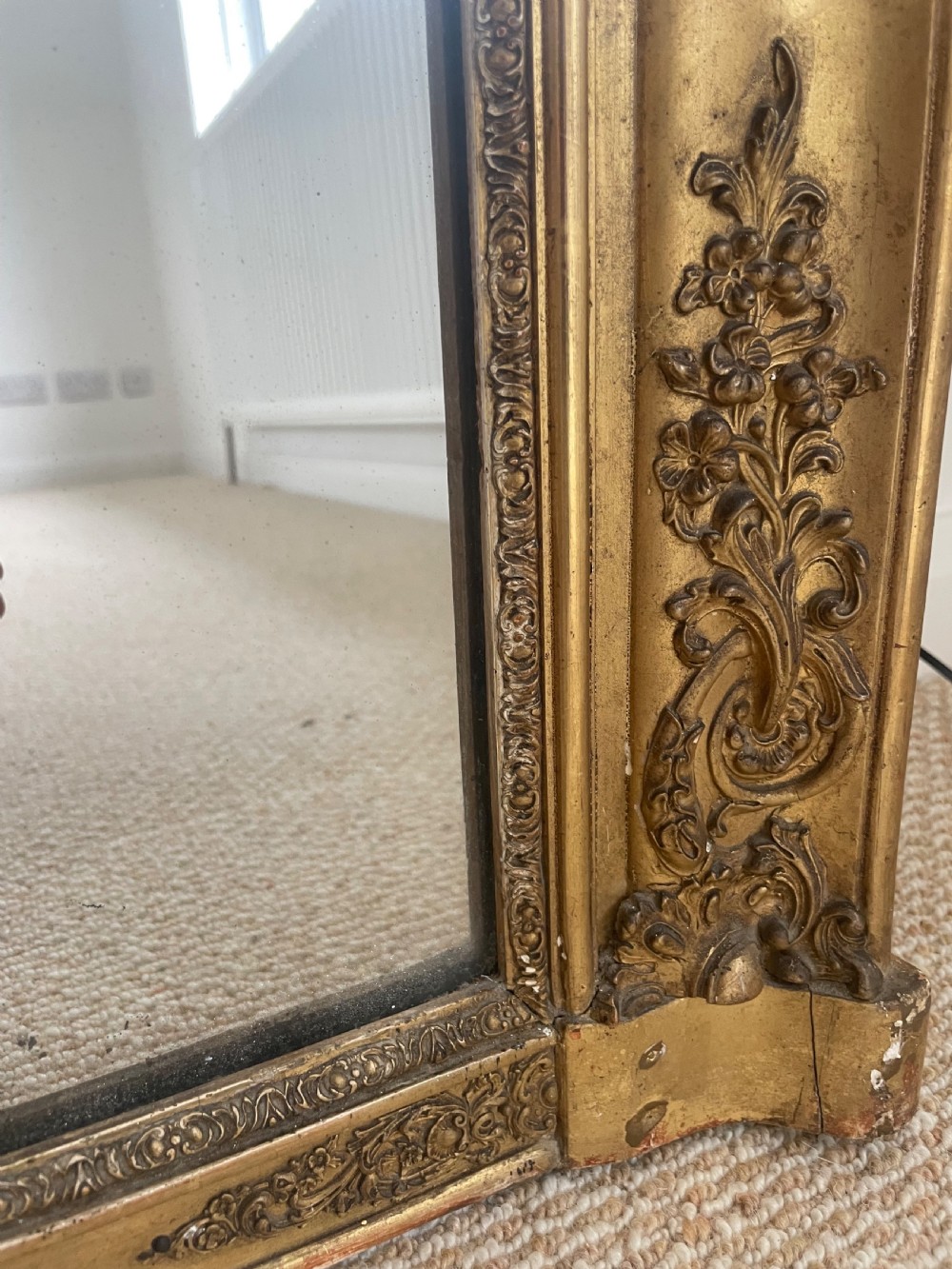 A large rectangular antique overmantel mirror featuring a gilded frame embellished with gesso scrollwork, floral motifs, and a central cartouche top emblem. View VI
