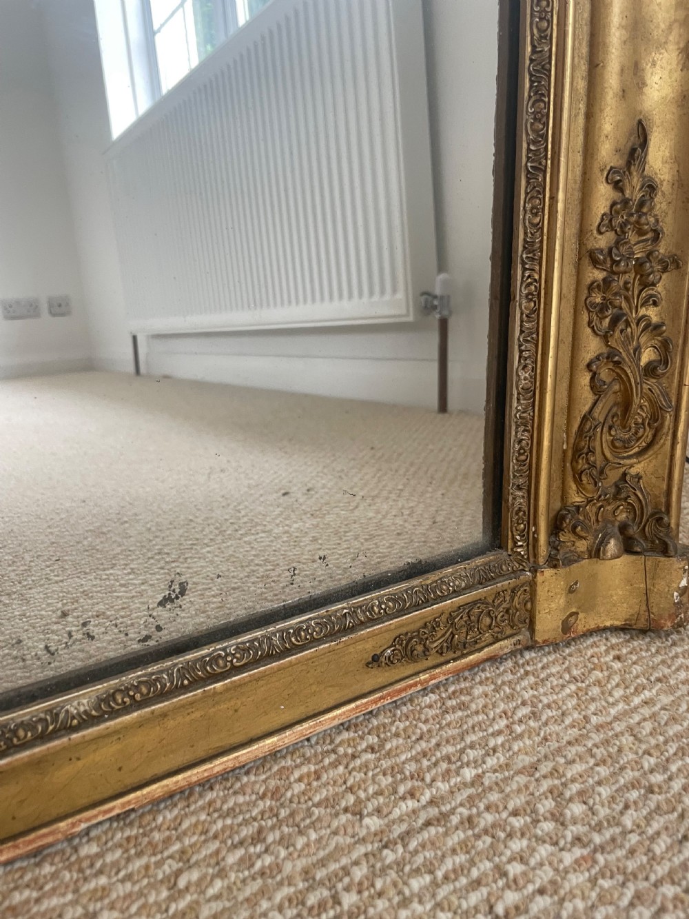 A large rectangular antique overmantel mirror featuring a gilded frame embellished with gesso scrollwork, floral motifs, and a central cartouche top emblem. View VII