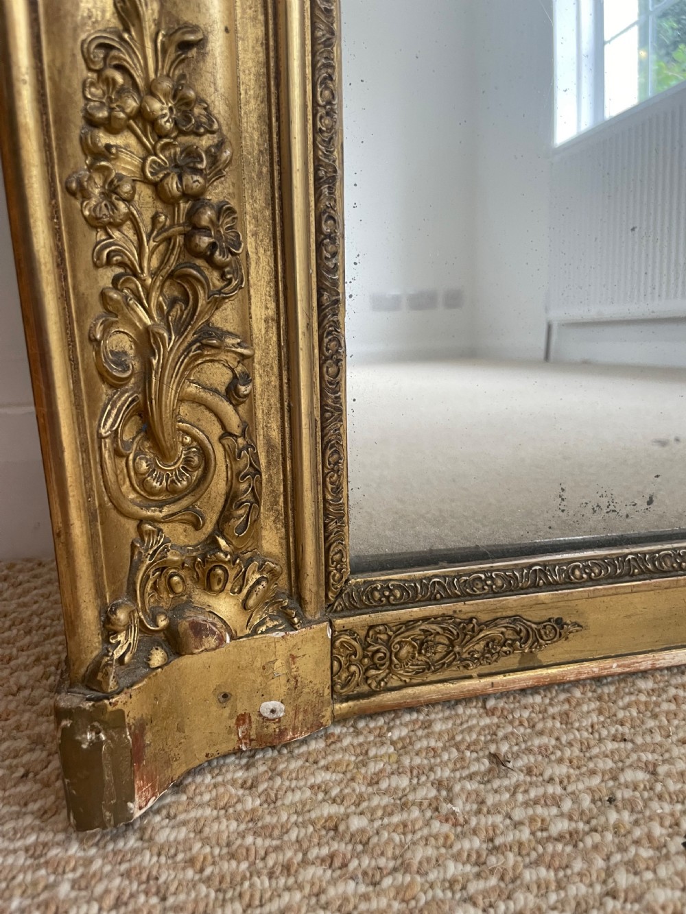 A large rectangular antique overmantel mirror featuring a gilded frame embellished with gesso scrollwork, floral motifs, and a central cartouche top emblem. View IX
