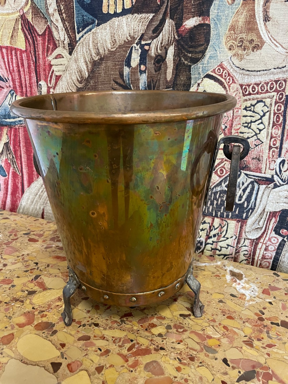 A tapered round copper container featuring riveted iron feet, a bottom band with visible rivets, and two side ring handles against a patterned textile backdrop. View II