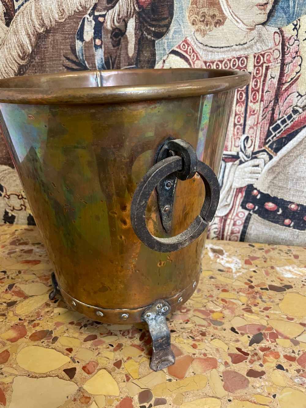 A tapered round copper container featuring riveted iron feet, a bottom band with visible rivets, and two side ring handles against a patterned textile backdrop. View III