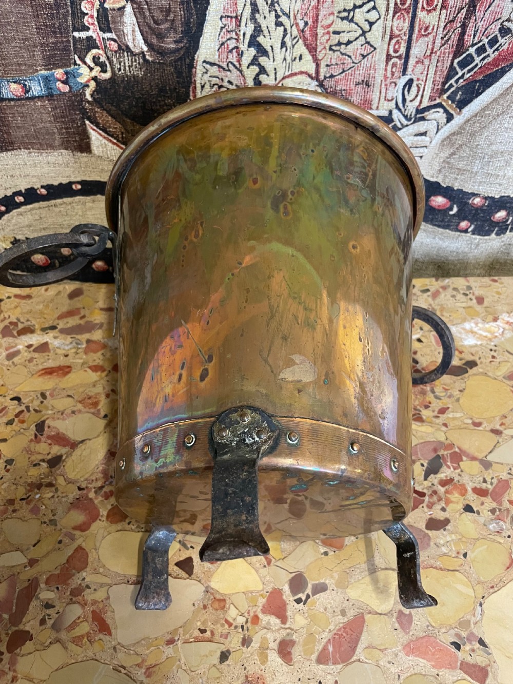 A tapered round copper container featuring riveted iron feet, a bottom band with visible rivets, and two side ring handles against a patterned textile backdrop. View IV