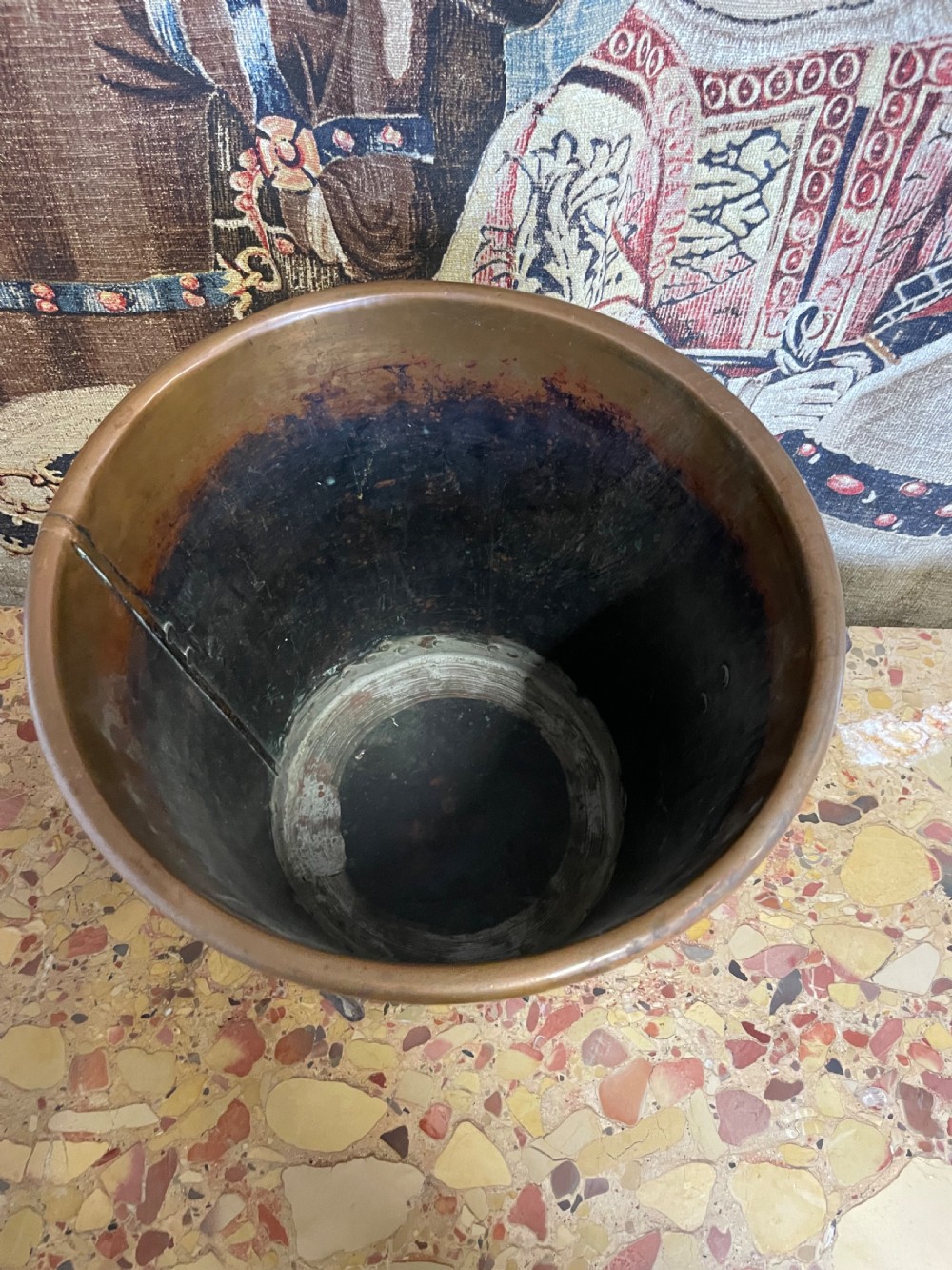 A tapered round copper container featuring riveted iron feet, a bottom band with visible rivets, and two side ring handles against a patterned textile backdrop. View V
