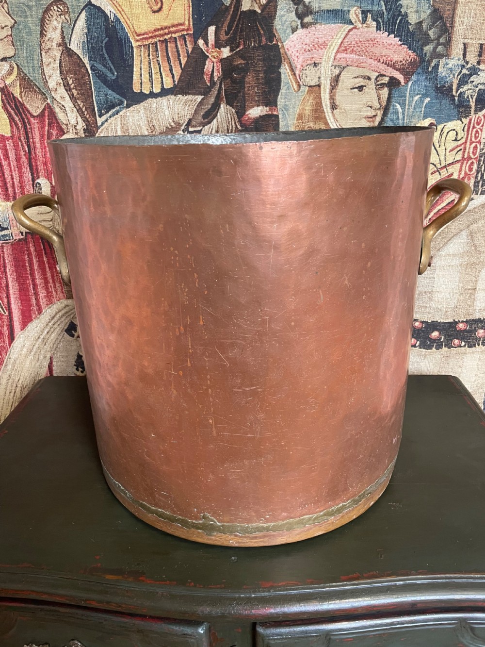 A large cylindrical copper log bin featuring two cast brass side handles and a hammered metal finish.