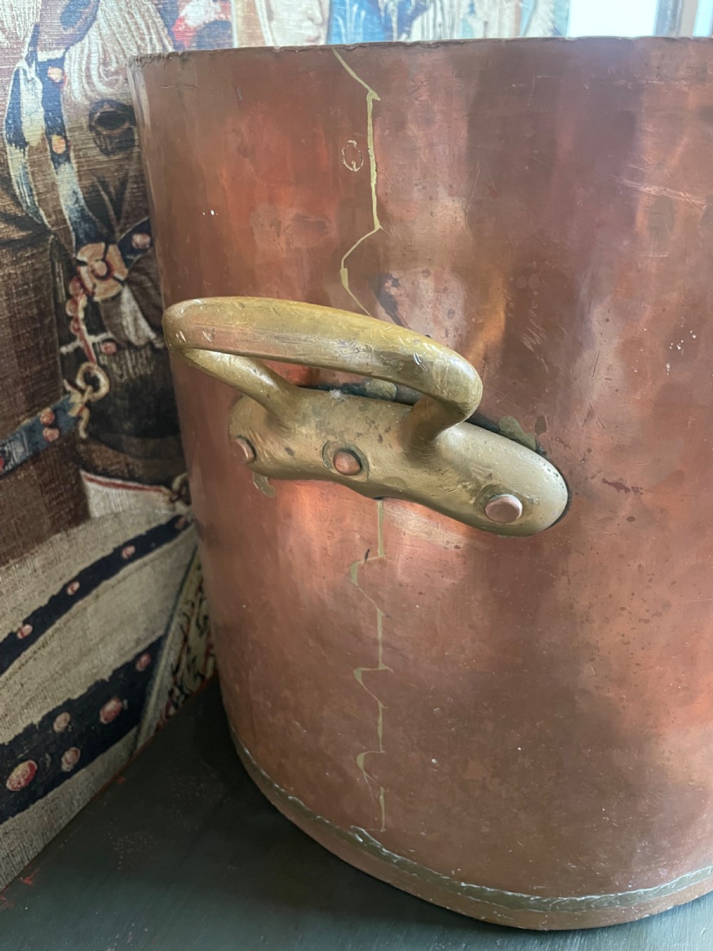 A large cylindrical copper log bin featuring two cast brass side handles and a hammered metal finish. View VI