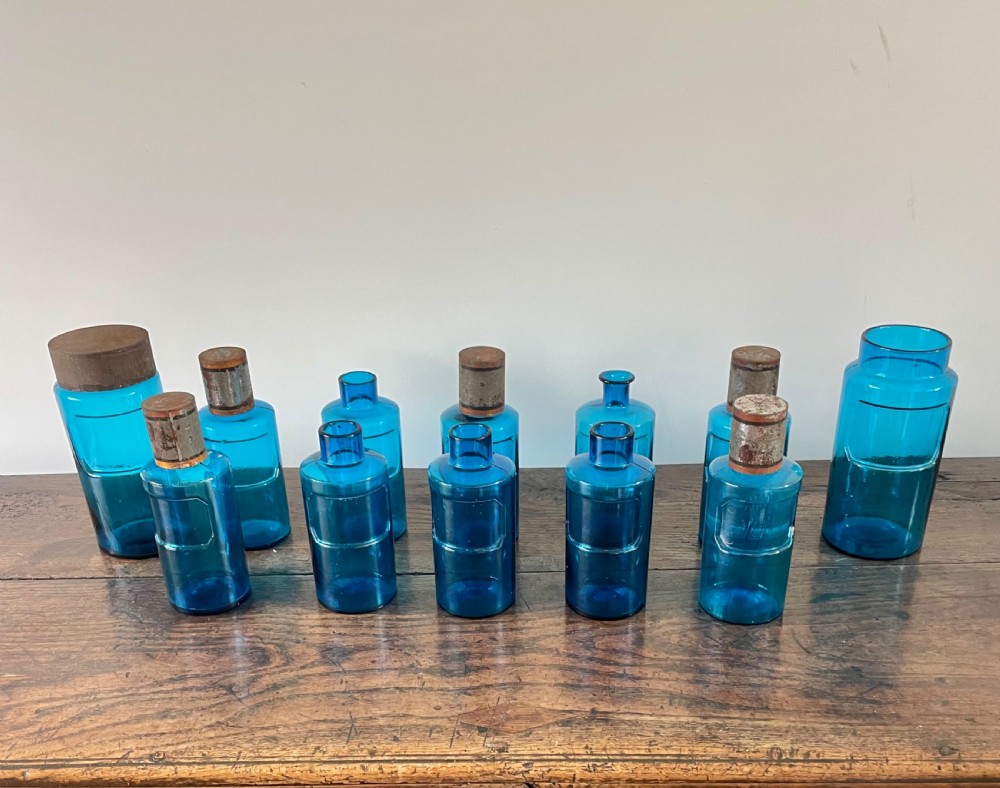 A collection of twelve cylindrical blue glass apothecary bottles with fitted metal caps displayed on a rustic timber surface.