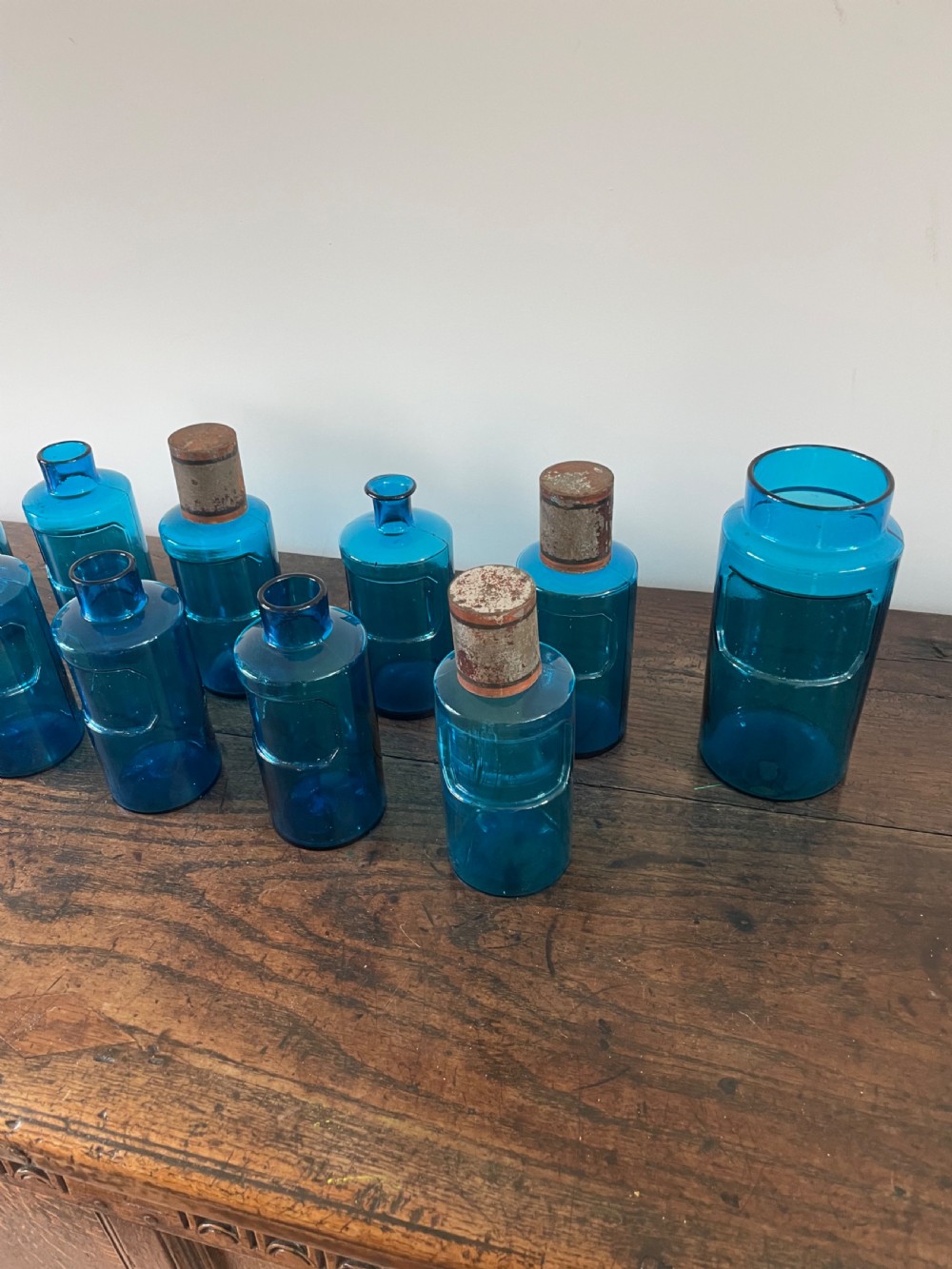 A collection of twelve cylindrical blue glass apothecary bottles with fitted metal caps displayed on a rustic timber surface. View II
