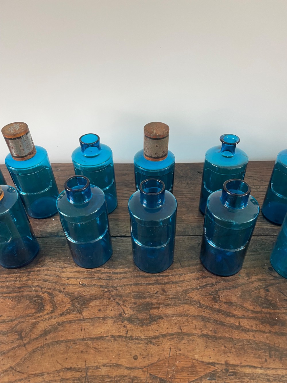 A collection of twelve cylindrical blue glass apothecary bottles with fitted metal caps displayed on a rustic timber surface. View III