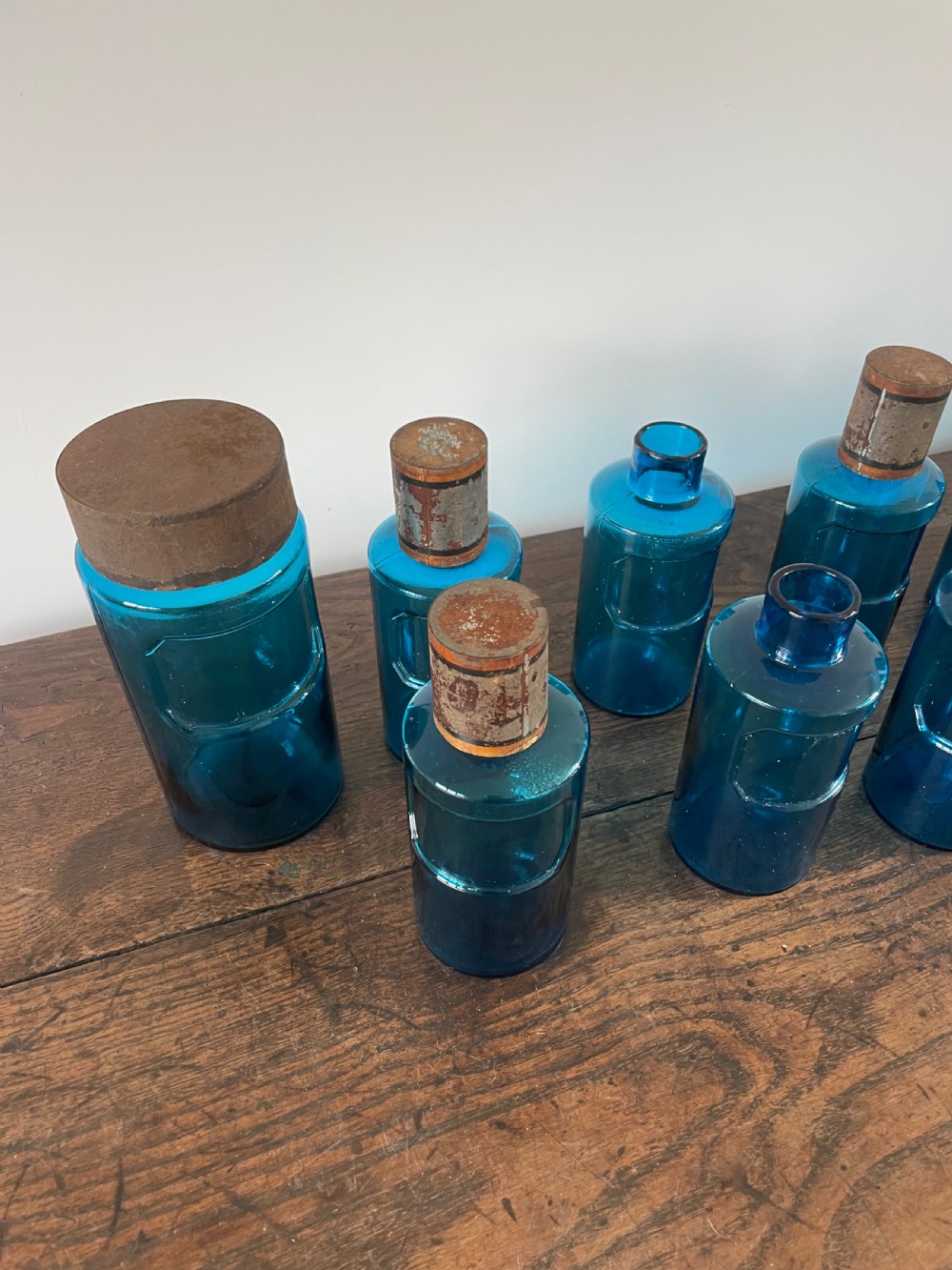 A collection of twelve cylindrical blue glass apothecary bottles with fitted metal caps displayed on a rustic timber surface. View IV