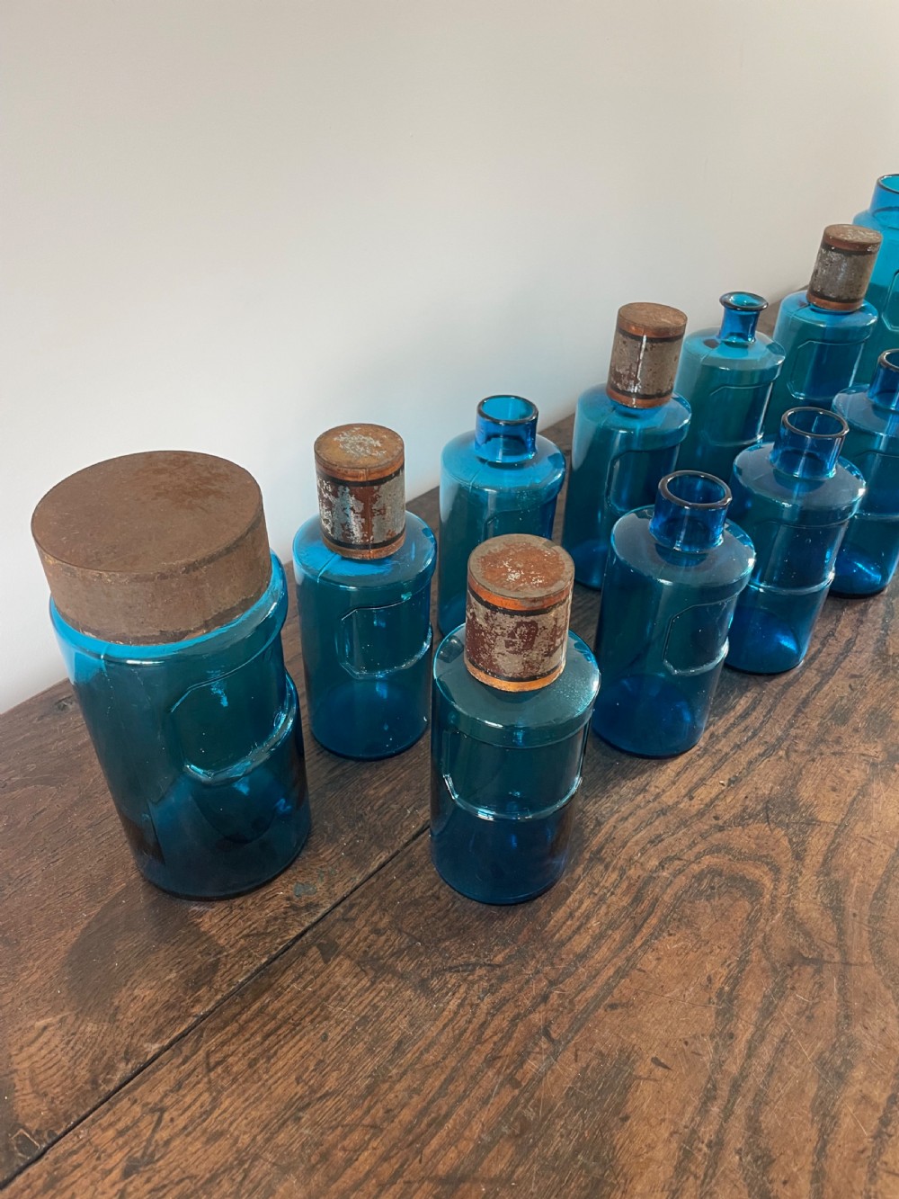 A collection of twelve cylindrical blue glass apothecary bottles with fitted metal caps displayed on a rustic timber surface. View V