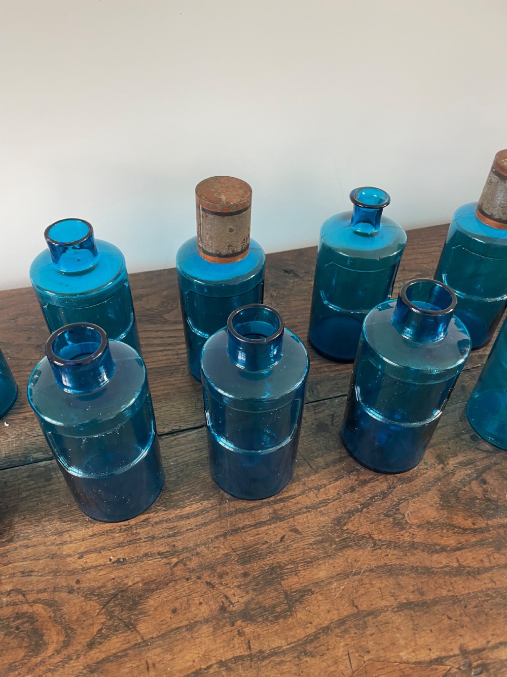 A collection of twelve cylindrical blue glass apothecary bottles with fitted metal caps displayed on a rustic timber surface. View VI