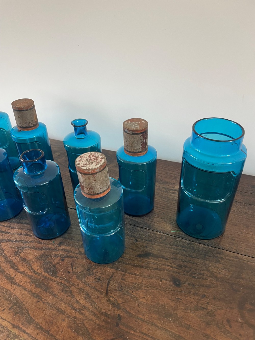 A collection of twelve cylindrical blue glass apothecary bottles with fitted metal caps displayed on a rustic timber surface. View VII