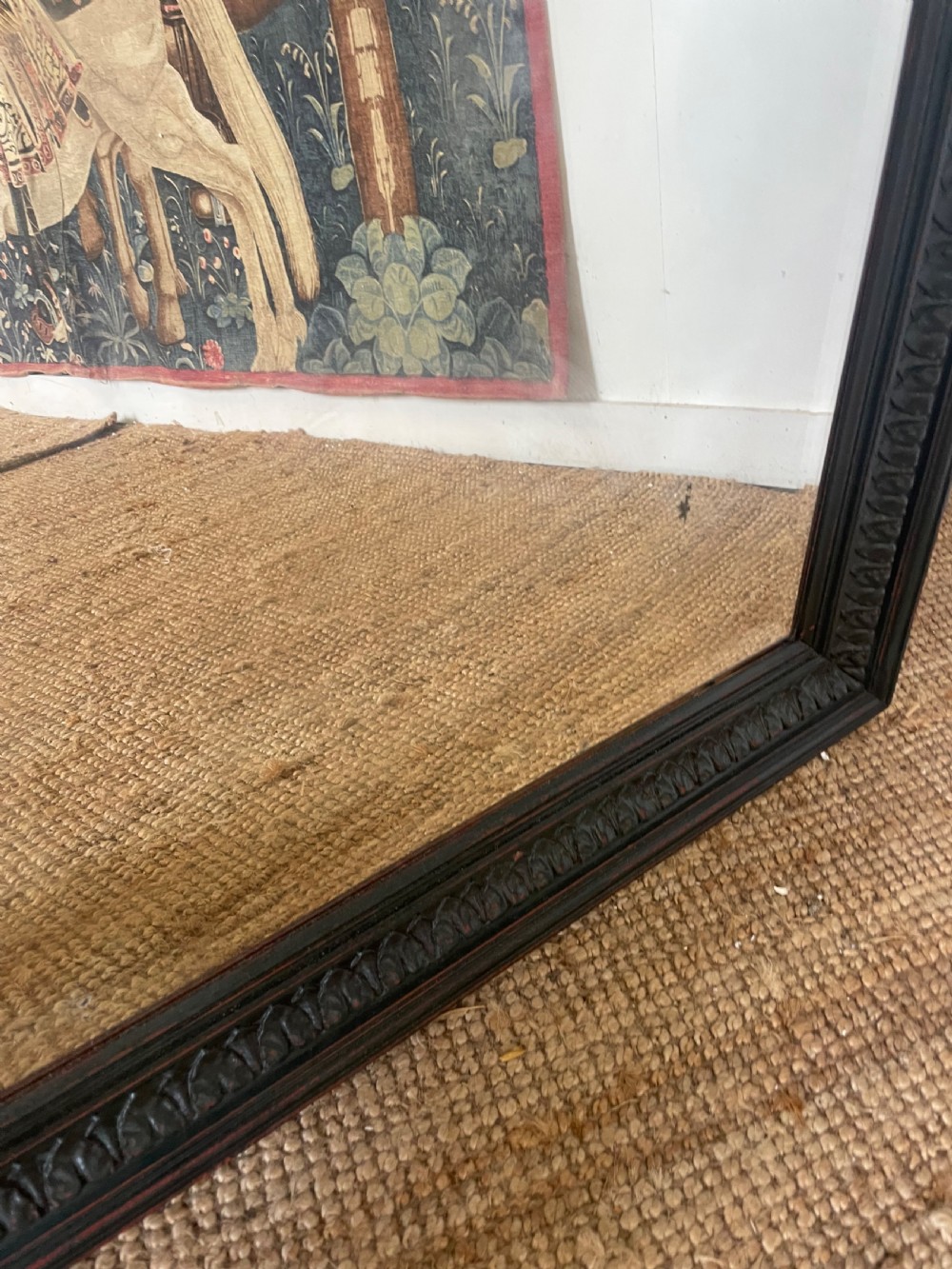 A large rectangular wall mirror with a dark, distressed carved wooden frame resting against a white wall. View VI