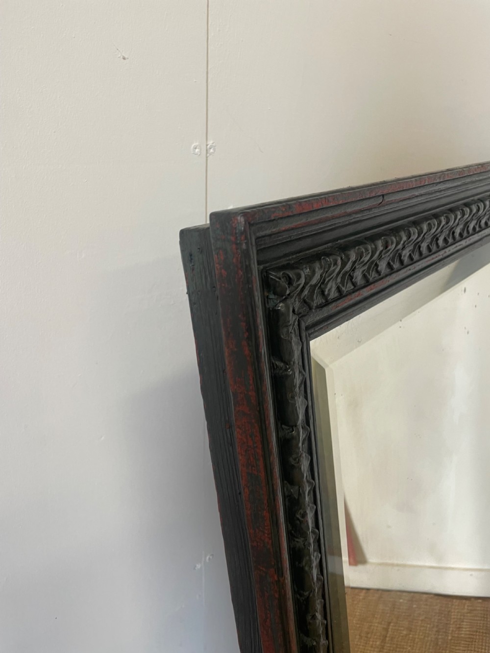 A large rectangular wall mirror with a dark, distressed carved wooden frame resting against a white wall. View VIII