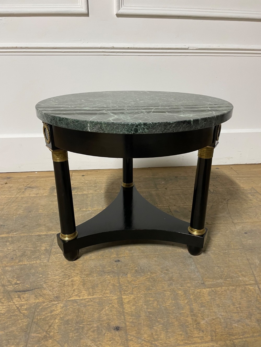 A circular Empire style table featuring a polished green marble top, ebonised column legs with gilt brass mounts, and a concave triform base.