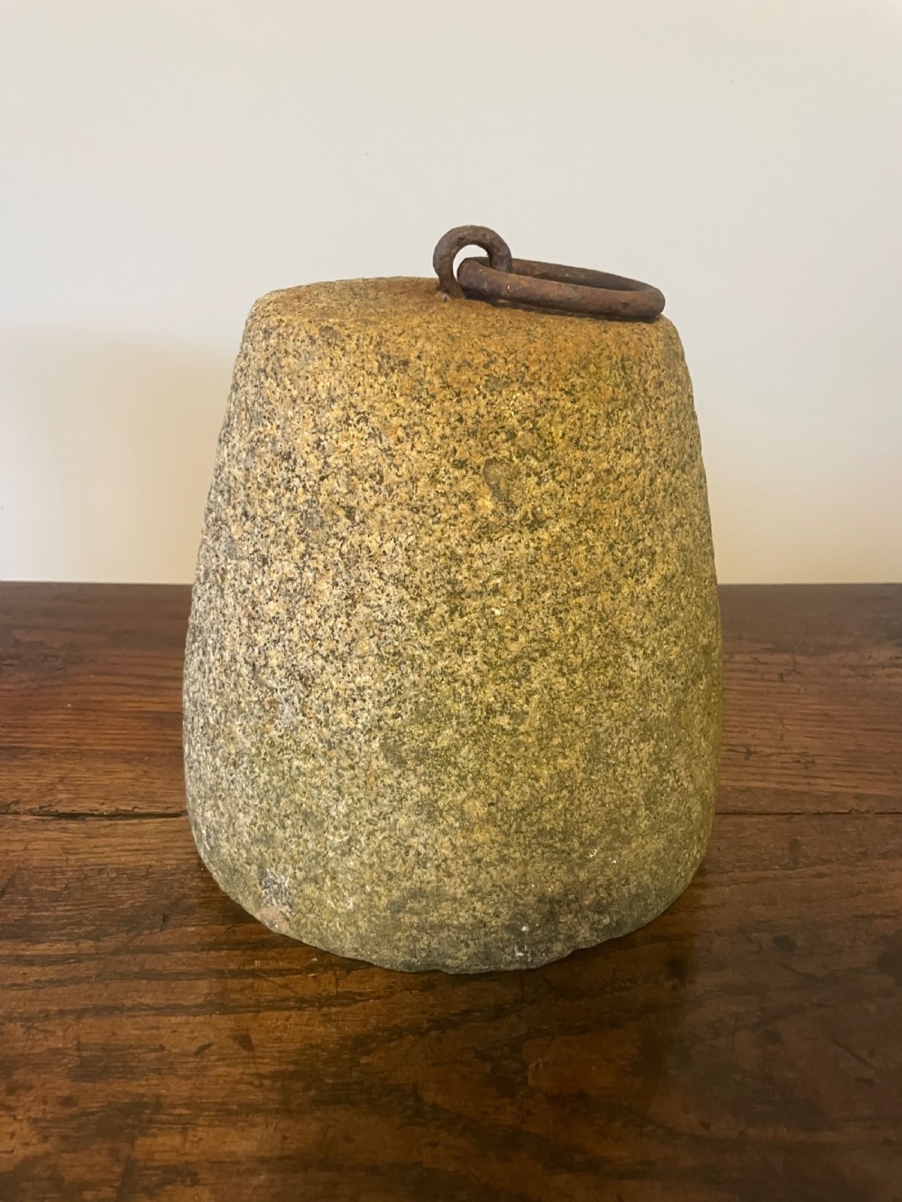 A weathered, tapered granite farm weight with an embedded rusted iron ring on top, resting on a dark wooden surface.