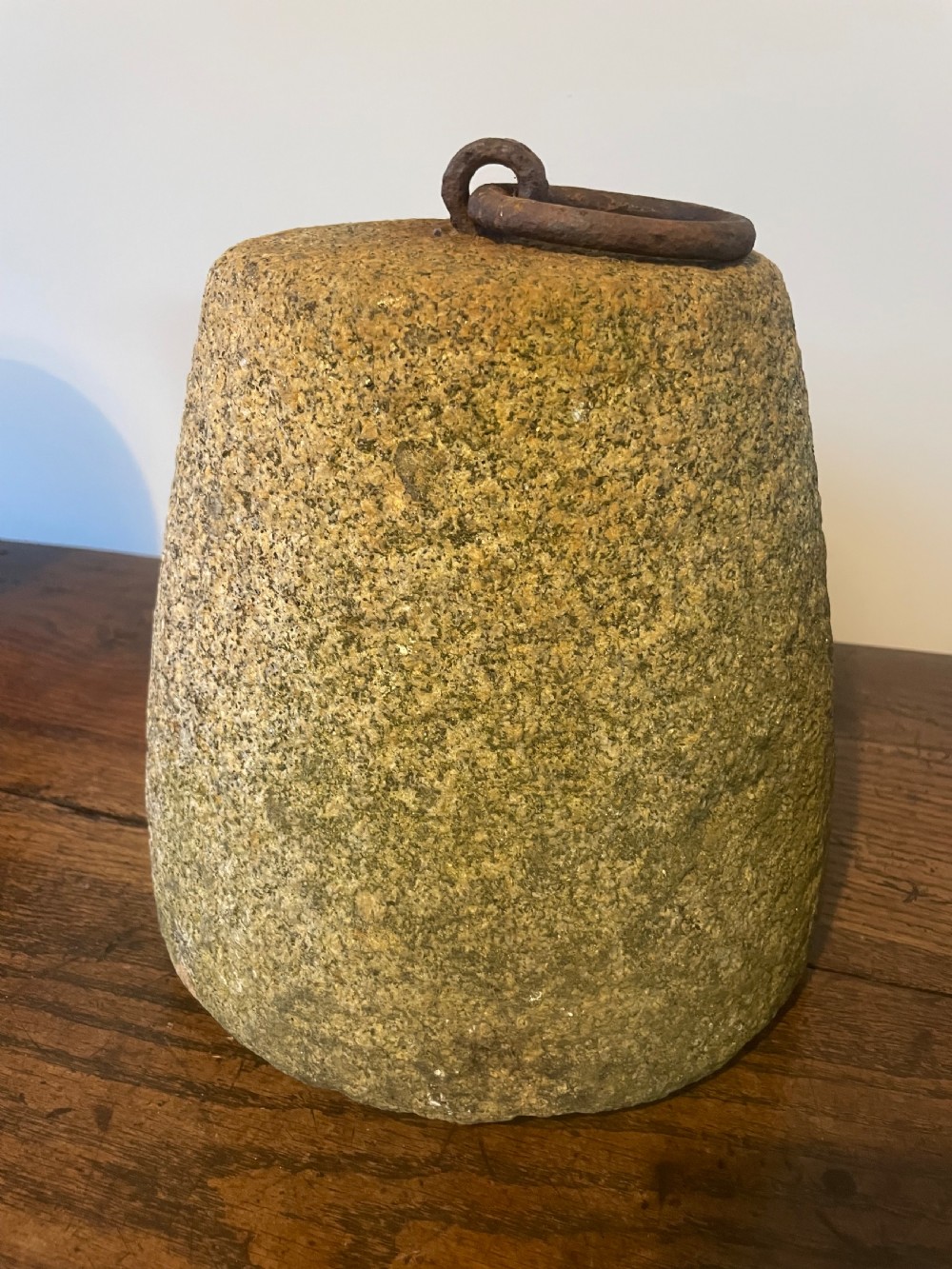 A weathered, tapered granite farm weight with an embedded rusted iron ring on top, resting on a dark wooden surface. View II