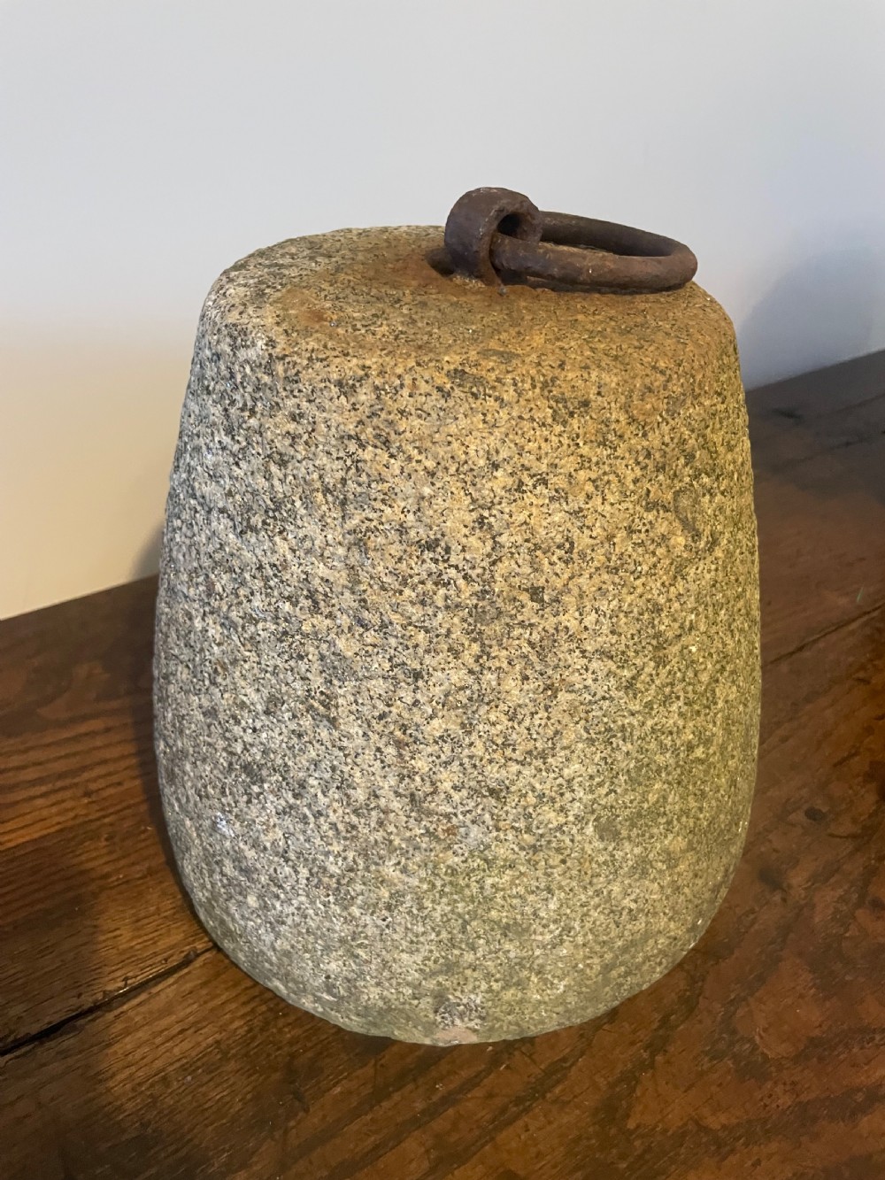 A weathered, tapered granite farm weight with an embedded rusted iron ring on top, resting on a dark wooden surface. View III