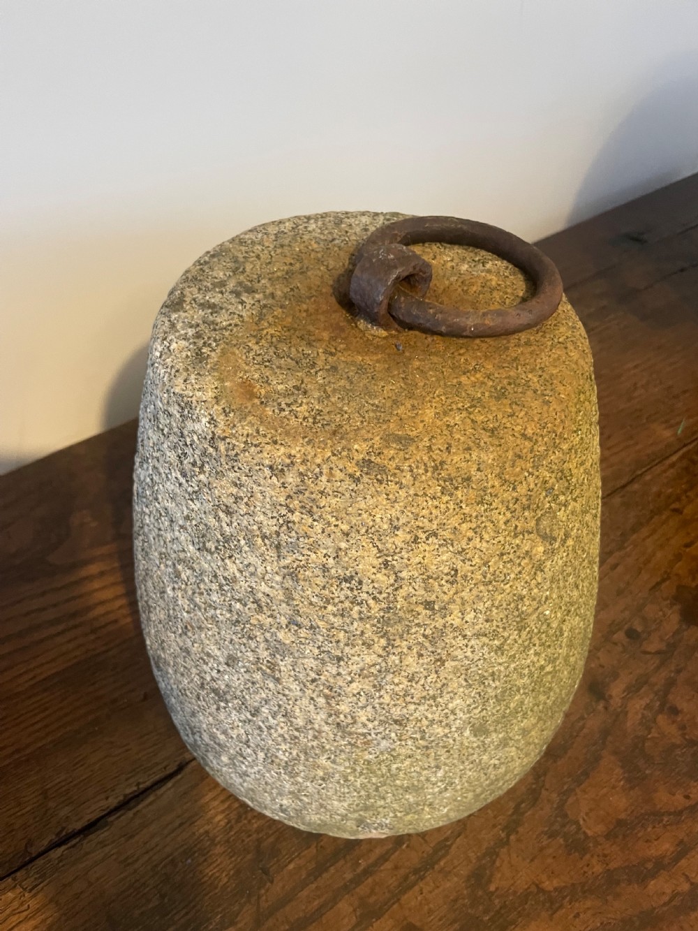 A weathered, tapered granite farm weight with an embedded rusted iron ring on top, resting on a dark wooden surface. View IV