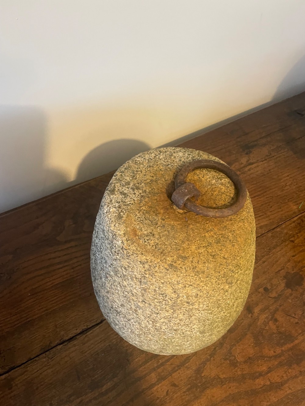 A weathered, tapered granite farm weight with an embedded rusted iron ring on top, resting on a dark wooden surface. View V